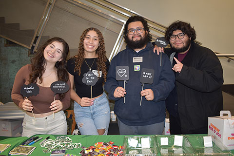 Four students stand behind a table holding handmade signs about being first generation college students.