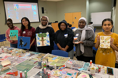 A group of first generation students stand behind a table full of arts and crafts supplies holding up their artwork.