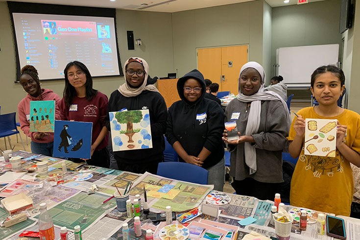 A group of students stand in a row holding paintings that they have created.