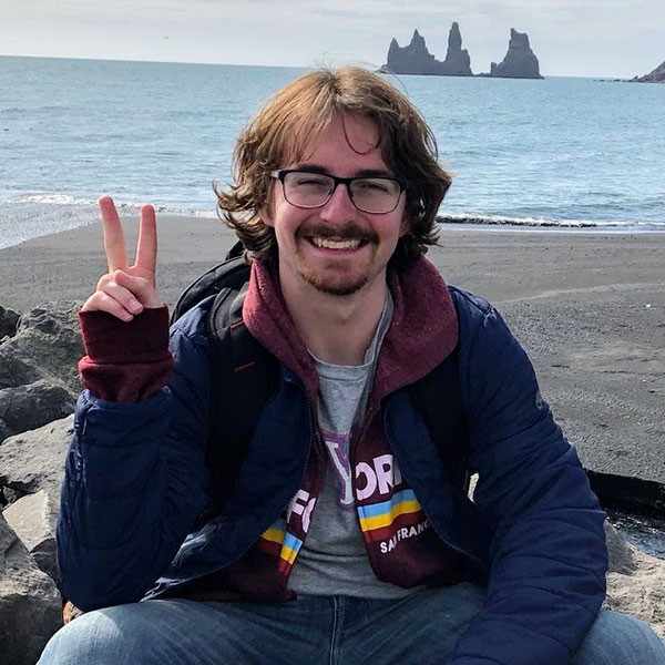 Max, holding up a peace sign, sits on rocks in front of the ocean