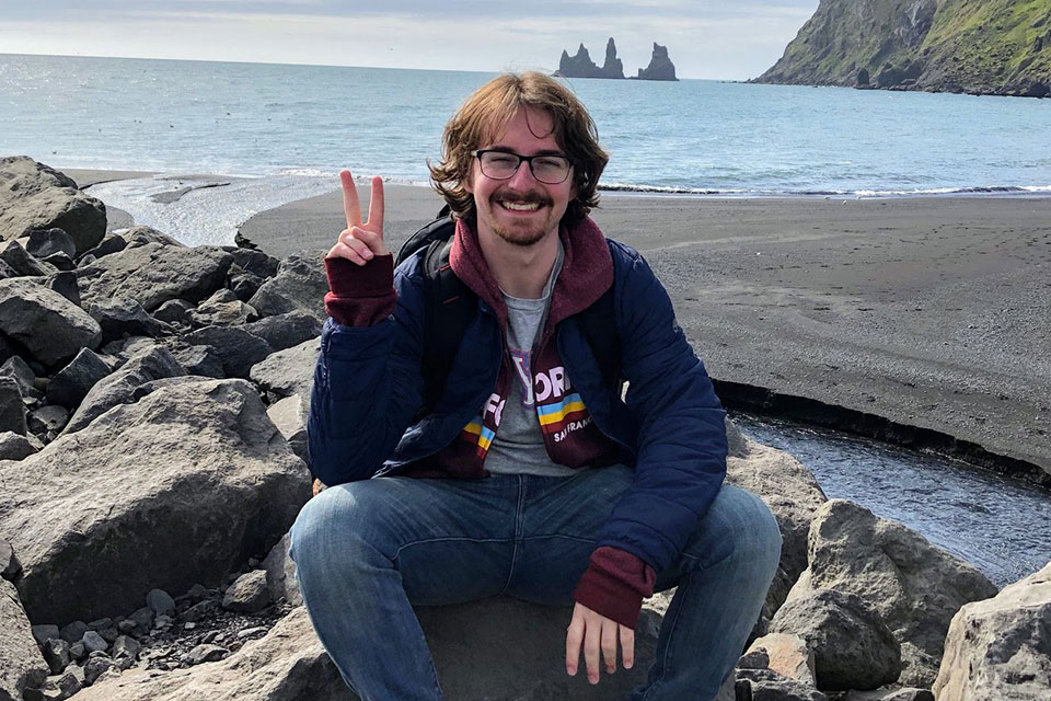 Max, holding up a peace sign, sits on the rocks in front of the ocean