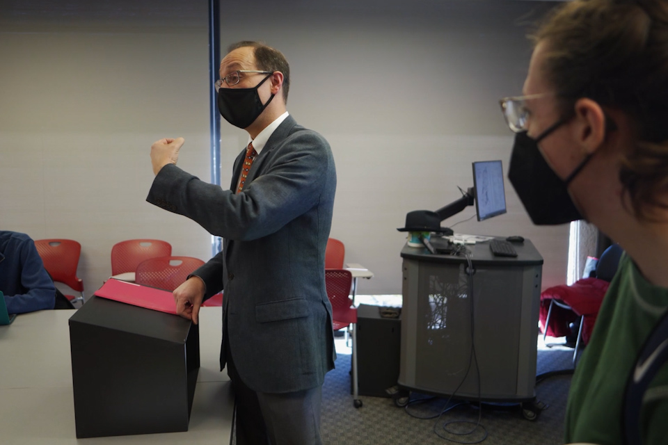 Govind Sreenivasan and a student standing at the front of a classroom. Both are wearing masks.