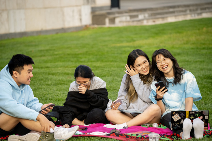 Four students are sitting on a blanket on a lawn. The two students on the right are smiling at the camera. 