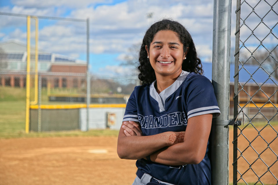 Rani smiling at the camera, wearing a Brandeis athletics jersey, standing at the edge of the softball field