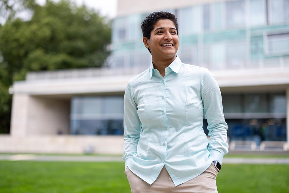 Rani smiling at the camera, wearing a Brandeis athletics jersey, standing at the edge of the softball field