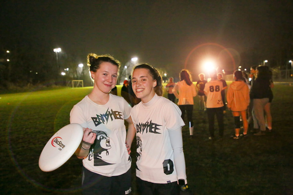 Eve and Allie smile at the camera. Eve is holding a frisbee and they are standing on a lit field at night.