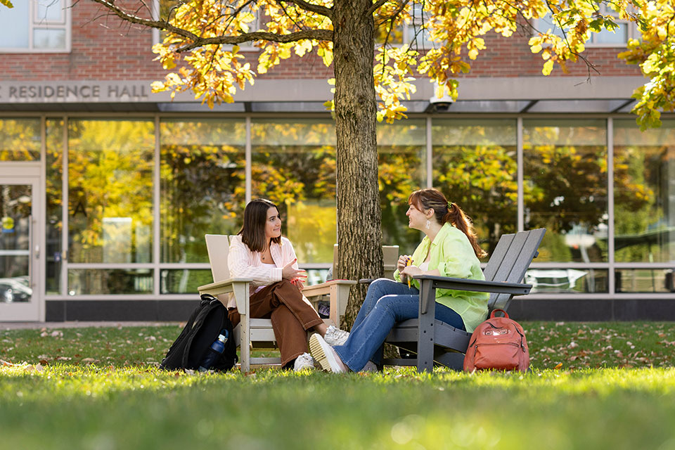 Two students sit together outside in chairs on a sunny day talking