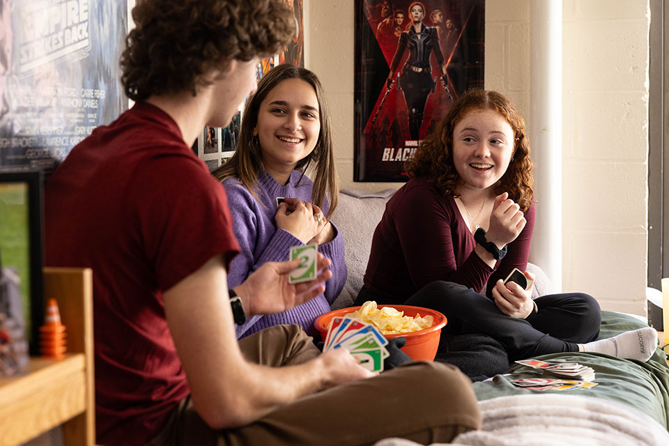 Students sitting on dorm room bed playing a card game and laughing