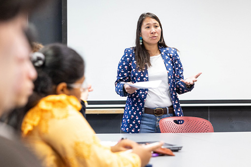 A professor stands at the end of a table in front of a white board