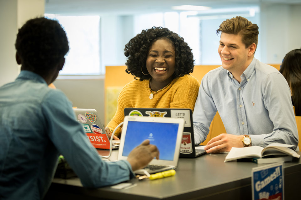 Three students sit laughing at a table in the library