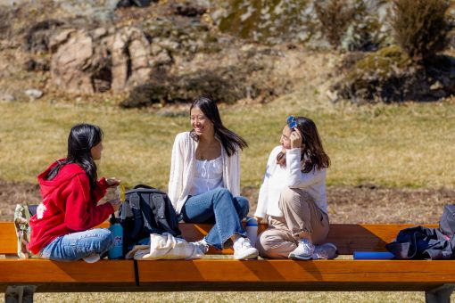 Three students sit on a bench on campus