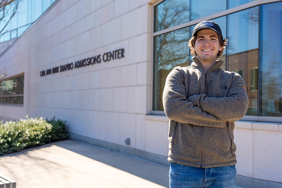 Ayden smiling at the camera with arms crossed. He is standing in front of the admissions center.