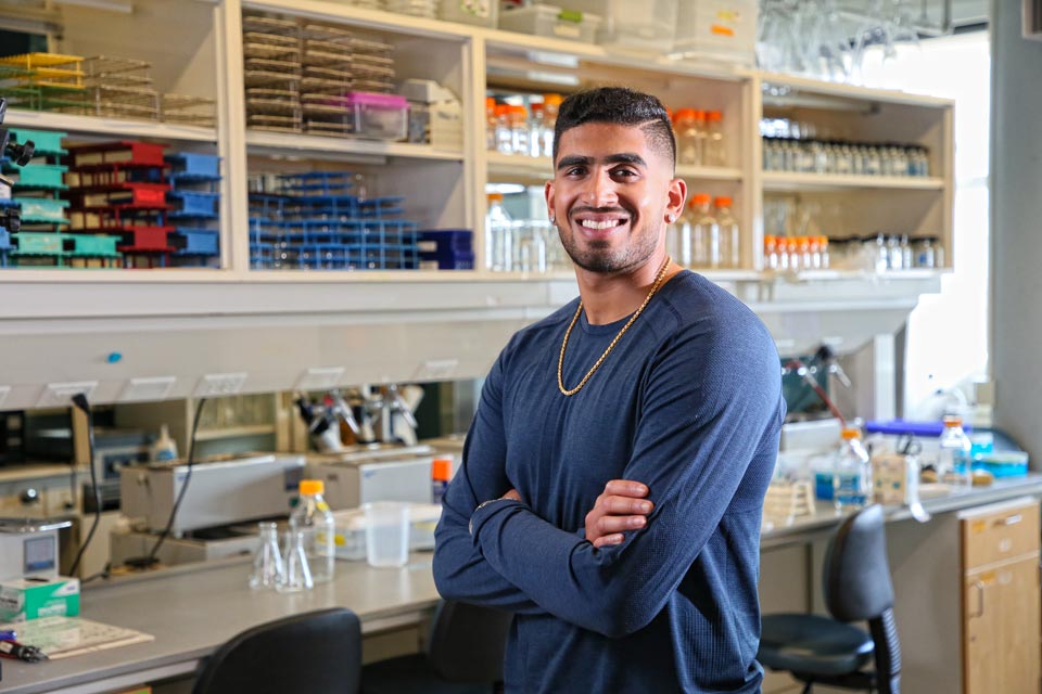 Aneesh smiling at the camera, standing in a science lab with shelves of equipment like bottles and beakers