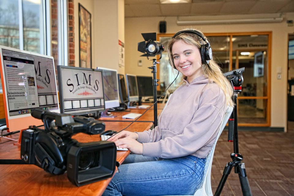 Haley smiling at the camera. She is wearing headphones and seated at a desk in the media studio with a video camera and a computer.