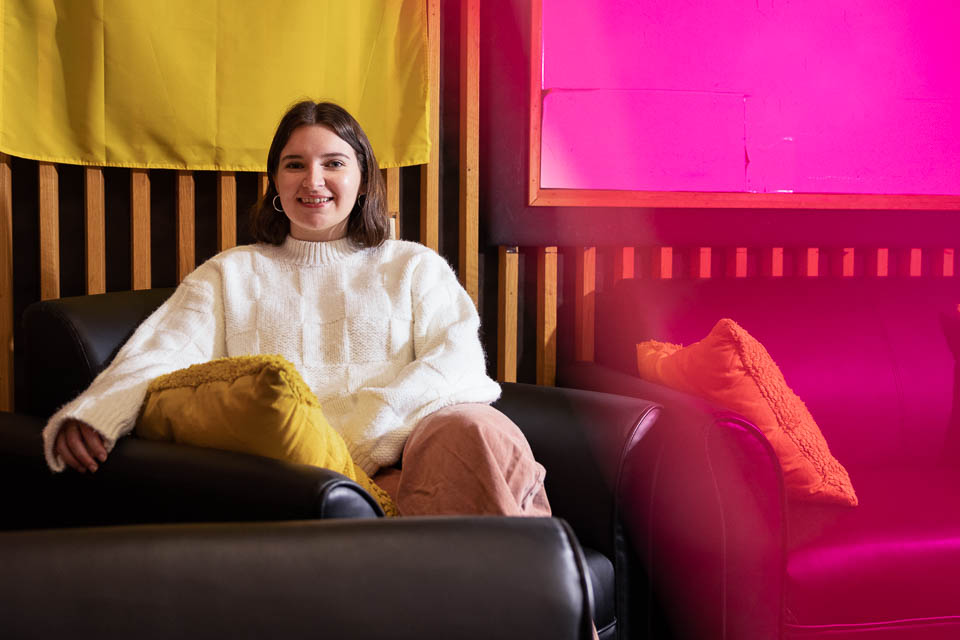 Kristianna sits in an arm chair with a yellow pillow. There is a yellow flag hanging in the background and half of the photo has a pink overlay.