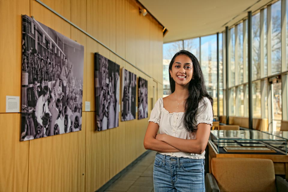 Nina smiling at the camera, standing in the lobby of the admissions center next to historical black and white Brandeis photos hanging on the wall.