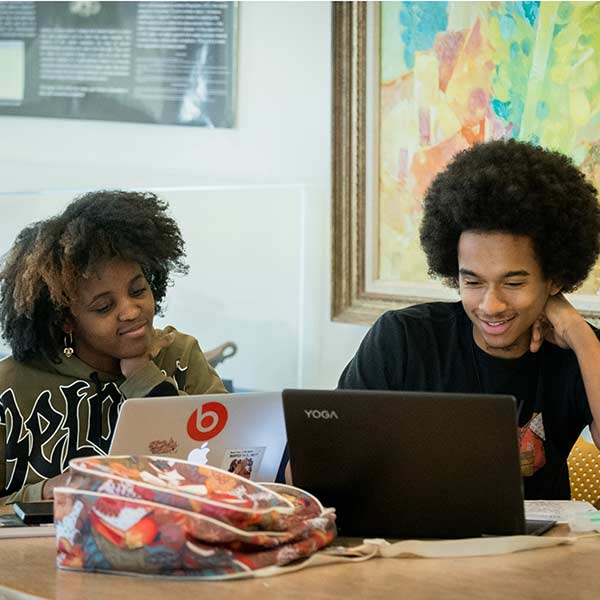 Two students sit next to each other while looking at laptops