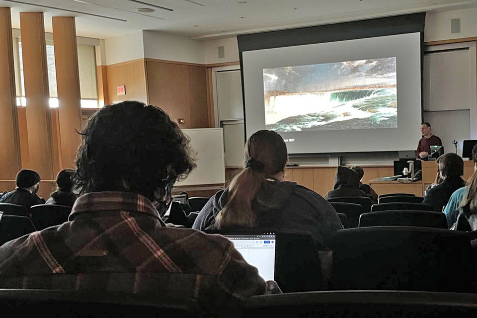 The view from Madison's seat in a classroom. The professor is standing at the front, behind a podium and a piece of artwork depicting waterfalls is project on a large screen.