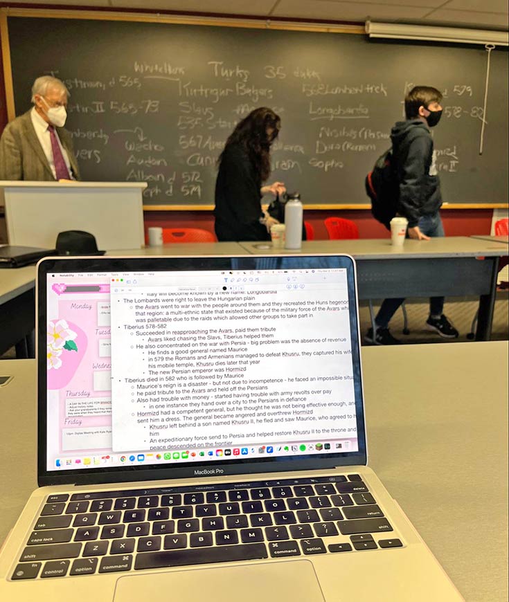 A few of a text document on Madison's laptop. In the background are two students and the professor in front of a chalkboard with writing.