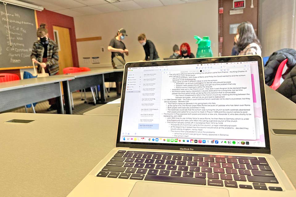 A view of a text document on Madisons laptop. In the background are other students around tables put together in a rectangle.