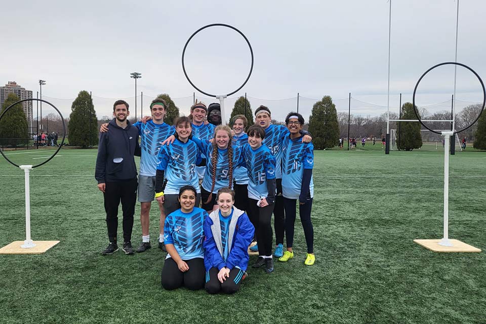 Marcus, his teammates and coach posing standing together on the quidditch field. They are wearing blue team shirts and there are circular goals around them