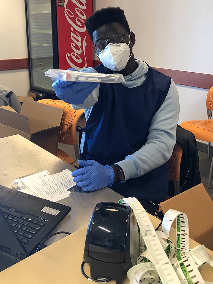 Marcus sitting in a chair, wearing a mask and holding a COVID test with his arm out toward the camera