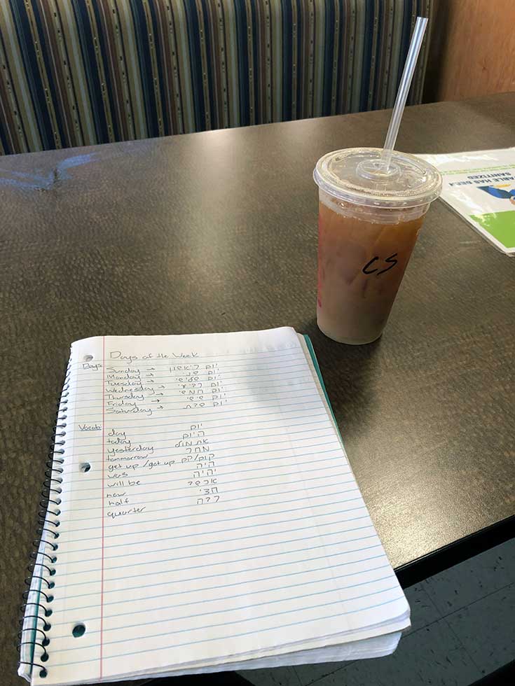 A spiral notebook with Hebrew writing on a table with a plastic cup with iced coffee