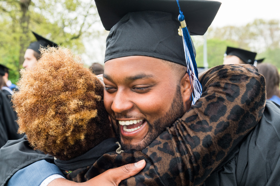 A male student smiling in cap and gown hugging his mother