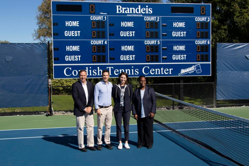 From left to right, President Ron Liebowitz, Jon Cordish, Melissa Fishman Cordish, Lauren Haynie, and Director of Athletics stand in front of the new Brandeis Cordish Tennis Center tennis scoreboard