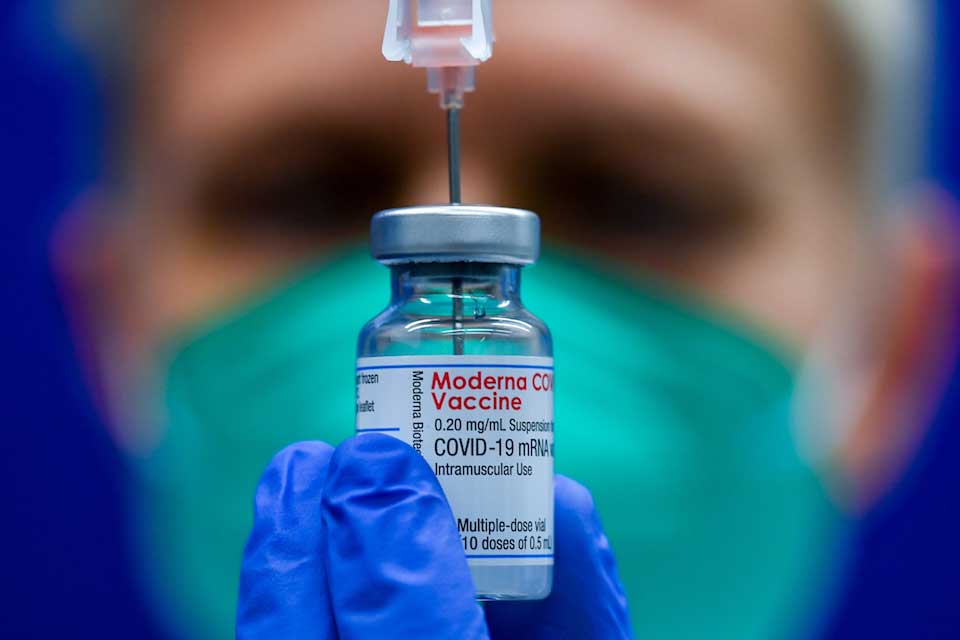 Researcher in background with close up of Moderna COVID-19 vaccine in foreground.