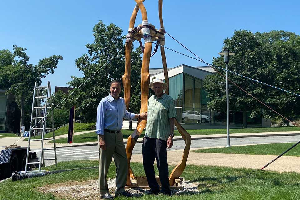 President Ron Liebowitz and Harold Grinspoon stand on Brandeis campus by wooden sculpture.