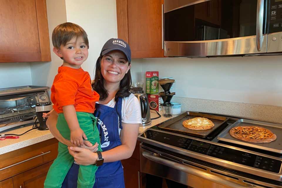 Monica LaBoskey holding her son and posing next to the stove with two pizzas on top.