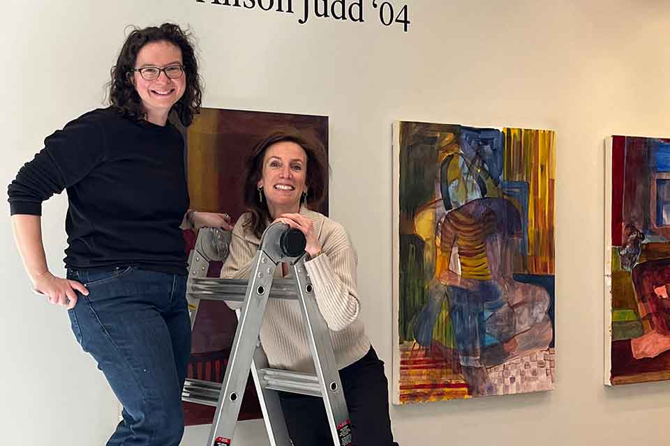 Alison Judd and Andrea Soloway standing on a ladder to install art.