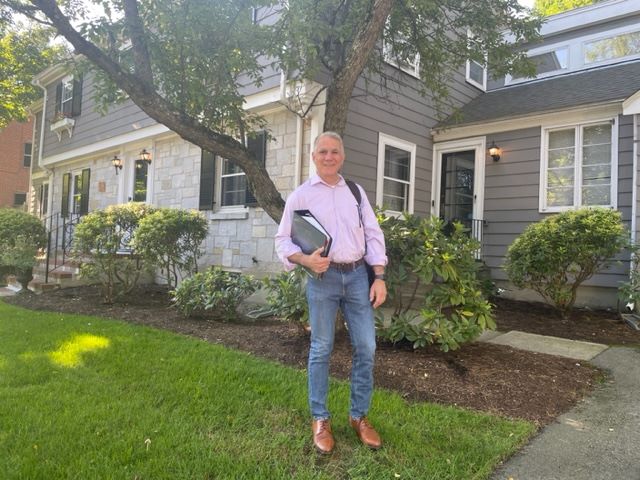 Jeffrey, a white man with grey hair, stands in front of a house with books and backpack