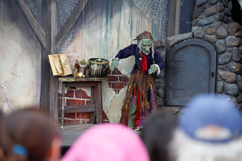 A witch marionette in front of an oven cauldron next to a worn table where her cauldron, elixirs, and book of spells sit, from a performance of Hansel and Gretel performed by Tanglewood Marionettes.