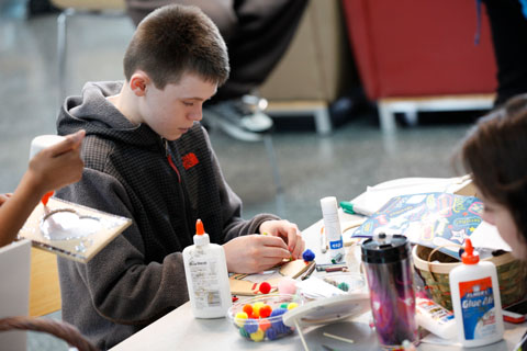 A young man crafting at a table full of art supplies including glue, crayons, small pom-poms, and stickers.