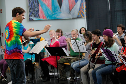 A conductor in a tie-dyed tee shirt in front of the wind section, conducting an orchestra.