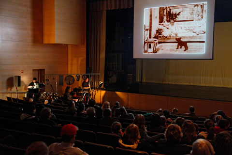 An audience watches a silent film on a movie screen accompanied by the Alloy Orchestra.