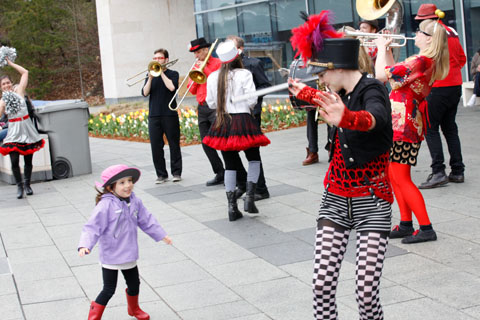 A young girl dancing with a Hungry March Band performer dressed playfully in red, black, and white.