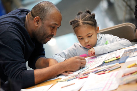 An adult helps a child at a table full of crayons pensively coloring intently.