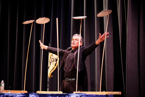 A performer with his hands spread wide in a black suit balances plates on tall sticks.