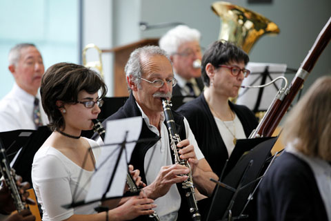 A man and a woman seated, playing clarinets with other musicians during a performance.