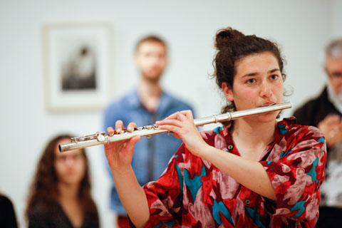 A flutist performing, with onlookers behind her.