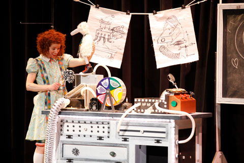 A red-haired woman performing on stage with an oversized lollipop and stuffed animal standing next to a desk of roughly assembled electronic looking devices under a clothes line with similarly looking sketches hung with binder clips.