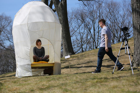 A photographer walks away from a camera tripod toward a rounded tent-like structure titled “Hive,” encompassing a student sitting with her legs folded.