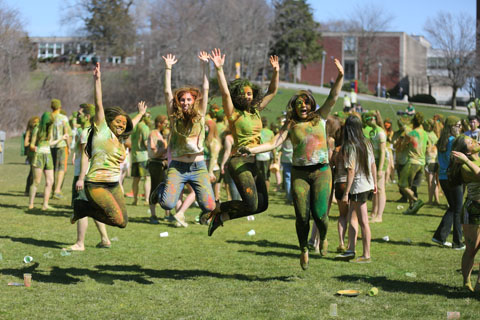 Students covered in colored powder jump in the air together for Holi.
