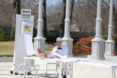 William Chambers dressed as a service attendant sitting and doing needlepoint for his live-performance installation titled “The Service Station.”