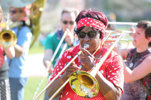 A colorfully-dressed woman from the Somerville School of Honk playing her slide trombone along with other horn players in the background.