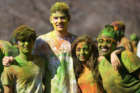 Students pose for the camera, smiling and covered in colorful powder during a Holi celebration.
