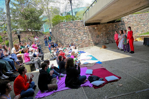 Student perform under a bridge outside, while the audience sits on the ground and looks on.
